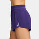 En kvinde er iført Nike Aeroswift 3" løbeshorts i Court Purple/White, der er fremstillet af bæredygtige materialer og har et hvidt Nike-logo på venstre side. Billedet er taget fra siden mod en almindelig hvid baggrund.