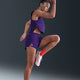 En kvinde iført Nike Aeroswift 3" løbeshorts i Court Purple/White, parret med hvide løbesko og orange sokker, løfter sit knæ og sin arm dynamisk mod en mørk baggrund.
