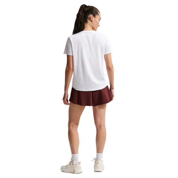 En kvinde står med ryggen til kameraet iført en hvid Nike Nk One Classic Df Ss Top (Dame), rødbrune shorts, hvide sokker og sneakers. Hendes mørke hår er sat op i en hestehale mod en almindelig hvid baggrund.