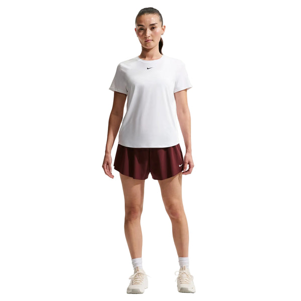 En kvinde står og kigger fremad iført en hvid/sort Nike Nk One Classic Df Ss Top og rødbrune Nike-shorts med hvide sokker og sportssko på en almindelig hvid baggrund.