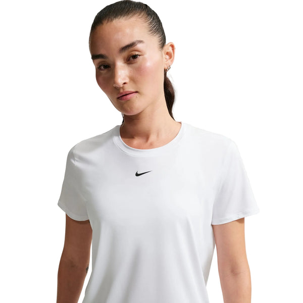 En kvinde med langt mørkt hår, der er bundet tilbage, er iført Nike Nk One Classic Df Ss Top, Dame, White/Black - en almindelig hvid T-shirt med et lille sort logo på brystet - på en hvid baggrund. Perfekt som trænings-T-shirt kvinder.