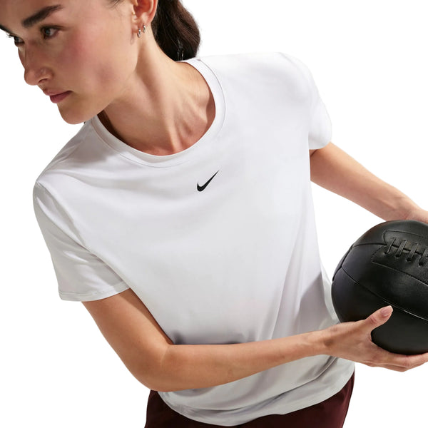 En person er iført en Nike Nk One Classic Df Ss Top, Dame, White/Black, og holder en sort medicinbold. Nike-logoet er synligt på trøjen mod en ren, hvid baggrund.