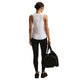 En kvinde kigger væk mod en almindelig hvid baggrund iført Nike Nk One Classic Df Tank (Dame, White/Black), sorte leggings og sko. Hun holder en sort sportstaske i venstre hånd og har håret bundet tilbage i en lav knold.