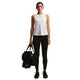 En kvinde kigger fremad i en Nike Nk One Classic Df Tank, Dame, White/Black og sorte leggings, mens hun holder en sort sportstaske og en vandflaske i venstre hånd. Hun har sorte sportssko på, alt sammen på en almindelig hvid baggrund.
