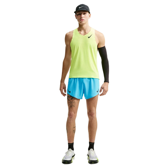 En person står med ansigtet vendt fremad iført en neongul tanktop, Nike AeroSwift 4" løbeshorts med indvendig underbuks i farven Lt Current Blue/Black, en sort armvarm, en sort kasket og sort-hvide løbesko; Nike-logoer er synlige.