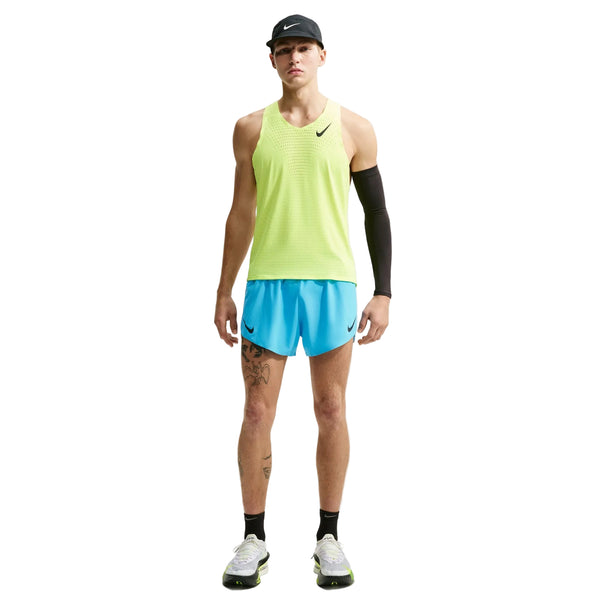 En person står med ansigtet vendt fremad iført en neongul tanktop, Nike AeroSwift 4" løbeshorts med indvendig underbuks i farven Lt Current Blue/Black, en sort armvarm, en sort kasket og sort-hvide løbesko; Nike-logoer er synlige.