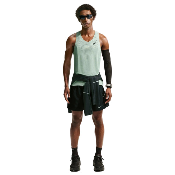 En person står med ansigtet fremad og er iført en grøn Nike AeroSwift Running Singlet, Herre, Steam/Black med Dri-FIT ADV-teknologi, sorte løbeshorts og sneakers, solbriller, et sort ærme og en sort jakke, der er bundet i taljen.