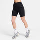 Iført Nikes One High-Waisted 8" Biker Shorts With Pockets i sort/sort, strømper og grå sneakers står en person på en almindelig lys baggrund. Kun underkroppen er synlig.