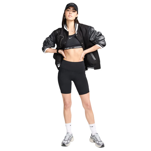 En person er iført sort kasket, sort universitetsjakke, sort sports-bh, Nike One High-Waisted 8" Biker Shorts With Pockets i Dame Black/Black, hvide strømper og grå sneakers med hænderne i jakkelommerne.