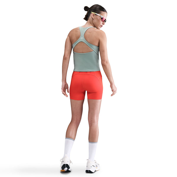 En person ser væk, iført en lysegrøn Nike Outdoor Bra Tank, Dame, Cannon med nøglehul i ryggen, knaldrøde, tætsiddende shorts, hvide sokker, sportssko og solbriller - ideel til udendørs træning. Almindelig hvid baggrund.