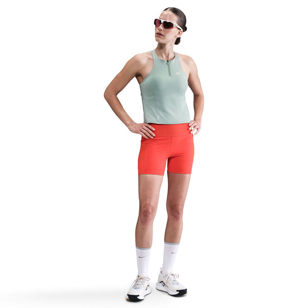 En kvinde iført Nikes Outdoor Bra Tank, Dame, Cannon med solbriller, røde sportsshorts, hvide knæstrømper og hvide sneakers står med hænderne på hofterne mod en almindelig hvid baggrund.