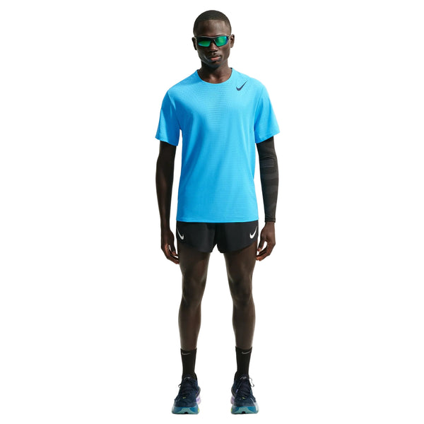 En person står med ansigtet vendt fremad iført en Nike AeroSwift løbetrøje med korte ærmer i farven Lt Current Blue/Black, sorte Nike-shorts, sorte sokker, løbesko, solbriller og en armvarmere foran en ensfarvet hvid baggrund.