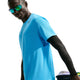 En person iført en lyseblå Nike AeroSwift kortærmet løbetrøje til mænd (Light Current Blue/Black), sorte armvarmere og reflekterende solbriller står foran en hvid baggrund og holder farverige hatte under den ene arm.