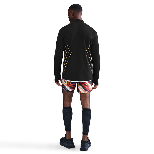 En person står med front mod kameraet iført en sort Nike Nk Df Jakob Hz langærmet skjorte til mænd, flerfarvede sportsshorts, sorte knæstrømper og sorte sportssko på en hvid baggrund.