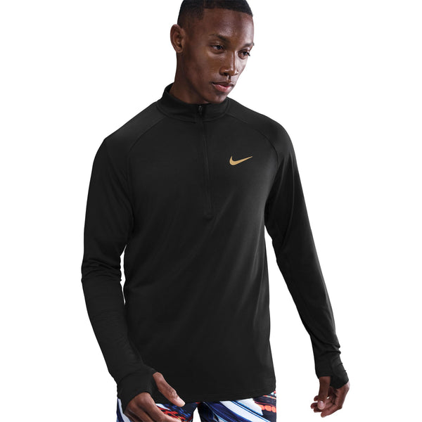 En mand bærer Nike Nk Df Jakob Hz-pulloveren i sort med guldfarvet swoosh og kvart lynlås sammen med lyse sportsshorts. Han står på en almindelig hvid baggrund.