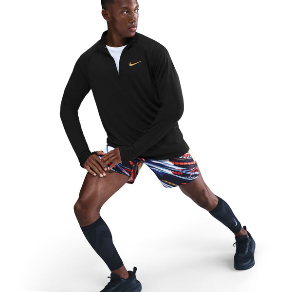 En mand iført en sort Nike Nk Df Jakob Hz langærmet sportstop, farvestrålende mønstrede shorts, sorte knæstrømper og sorte sneakers strækker benene mod en hvid baggrund.