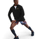 En mand iført en sort Nike Nk Df Jakob Hz langærmet sportstop, farvestrålende mønstrede shorts, sorte knæstrømper og sorte sneakers strækker benene mod en hvid baggrund.
