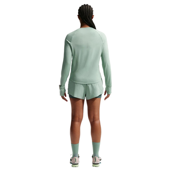 En person med flettet hår kigger væk fra kameraet iført en lysegrøn langærmet top, Nike Nk Swift Df Mr 2N1 Shrt til kvinder i Steam/Mineral Slate/Reflective Silver, strømper og sportssko på en almindelig hvid baggrund.