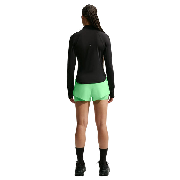 En person står med ryggen til og er iført en sort langærmet bluse, Nike Swift Mid-Rise 2-i-1-løbeshorts til kvinder i farven Illusion Green/Reflective Silver med Dri-FIT-teknologi, sorte sokker og sorte sko. Baggrunden er ensfarvet hvid. Personens hår er sat op i lange fletninger.