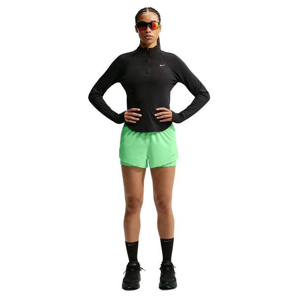 En person står med ansigtet vendt fremad og er iført en sort langærmet Nike-trøje, Swift Mid-Rise 2-i-1-løbeshorts til kvinder i farven Illusion Green/Reflective Silver fra Nike med Dri-FIT-teknologi, sorte sokker, sorte sneakers og rødtonede solbriller. Hænderne er lagt på hofterne, og håret er flettet.