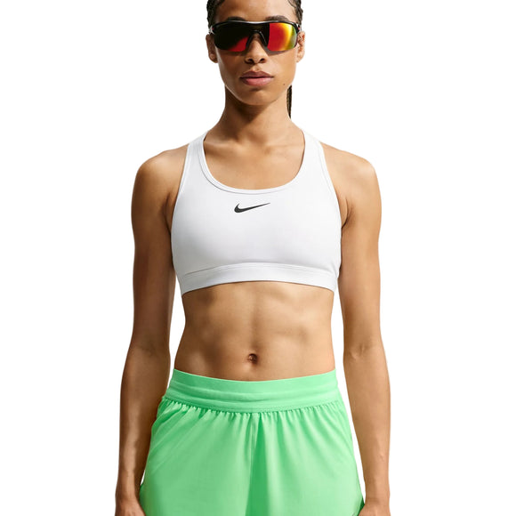 En person står med ansigtet vendt fremad foran en hvid baggrund og er iført en hvid Nike-sport-bh, Nike Swift Mid-Rise 2-i-1-løbeshorts til kvinder i farven Illusion Green/Reflective Silver med Dri-FIT-teknologi samt rødtonede sportsolbriller.