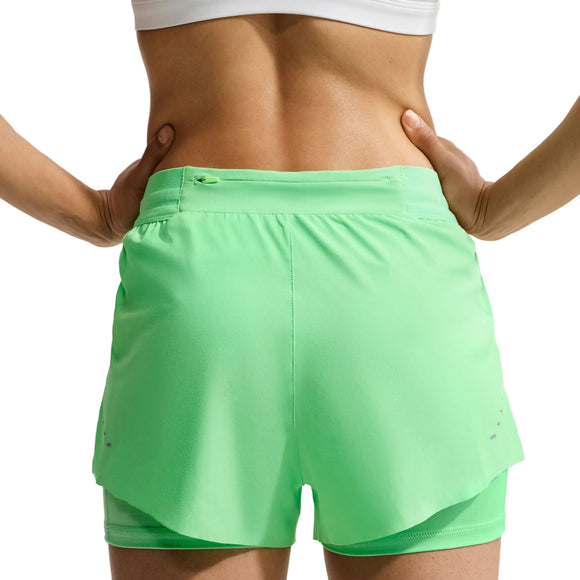 Der ses en person set bagfra med hænderne på hofterne, iført en hvid sports-BH og Nike Swift Mid-Rise 2-i-1-løbeshorts i damemodel, farven Illusion Green/Reflective Silver. Disse løbeshorts har et lagdelt design, Dri-FIT-teknologi til fugtregulering og en lille lomme med lynlås i linningen.