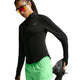 En person iført en sort Nike-trøje med lange ærmer, Swift Mid-Rise 2-i-1-løbeshorts til kvinder i farven Illusion Green/Reflective Silver fra Nike samt røde reflekterende solbriller strækker benet, mens vedkommende holder en sort løbesko i hånden.