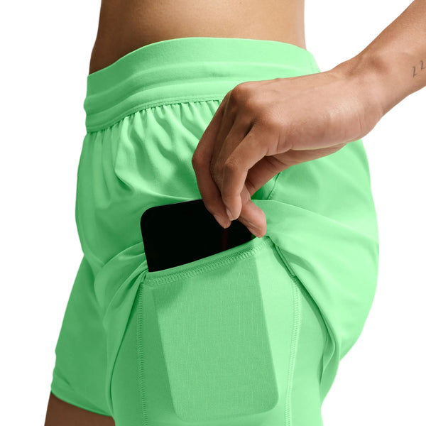 En person, der har Nike Swift Mid-Rise 2-i-1-løbeshorts til kvinder i farven Illusion Green/Reflective Silver med Dri-FIT-teknologi på, stikker en sort smartphone ned i en sidelomme. Kun personens overkrop og hånd er synlige på en hvid baggrund.