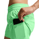 En person, der har Nike Swift Mid-Rise 2-i-1-løbeshorts til kvinder i farven Illusion Green/Reflective Silver med Dri-FIT-teknologi på, stikker en sort smartphone ned i en sidelomme. Kun personens overkrop og hånd er synlige på en hvid baggrund.