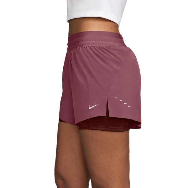 En person ses fra livet og ned i profil mod en ensfarvet baggrund, iført Nike Swift Mid-Rise 2-i-1-løbeshorts til kvinder i farvekombinationen Pink Smoke/Mystic Dates/Reflective Silv samt en hvid top. Shortsene er udstyret med Dri-FIT-teknologi, Nike-logoet og en lille slids i siden.