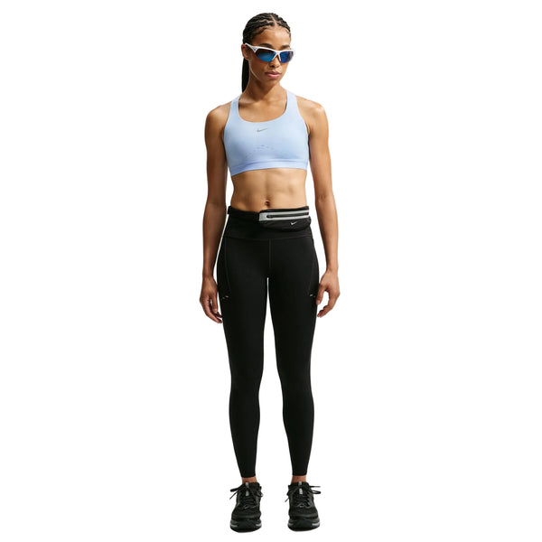 En person står med ansigtet vendt fremad og er iført en Nike High-Support Lightly Lined sports-BH til kvinder i farven Aluminium/Reflekterende Sølv, sorte leggings, sorte sneakers, en bæltetaske, blå solbriller og har langt flettet hår. Baggrunden er ensfarvet hvid.