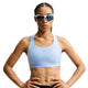 En kvinde iført en Nike High-Support Lightly Lined sports-BH til kvinder i farven Aluminium/Reflekterende Sølv står med hænderne i siden, vendt fremad mod en hvid baggrund, og har reflekterende solbriller på.