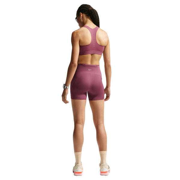 En kvinde med flettet hår står med ryggen til kameraet foran en ensfarvet hvid baggrund. Hun har en matchende Nike High-Support Lightly Lined sports-BH til kvinder i farven Pink Smoke/Reflective Silver på, kombineret med matchende shorts, hvide sokker, hvide og lyserøde sneakers, et ur og solbriller.