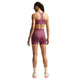 En kvinde med flettet hår står med ryggen til kameraet foran en ensfarvet hvid baggrund. Hun har en matchende Nike High-Support Lightly Lined sports-BH til kvinder i farven Pink Smoke/Reflective Silver på, kombineret med matchende shorts, hvide sokker, hvide og lyserøde sneakers, et ur og solbriller.