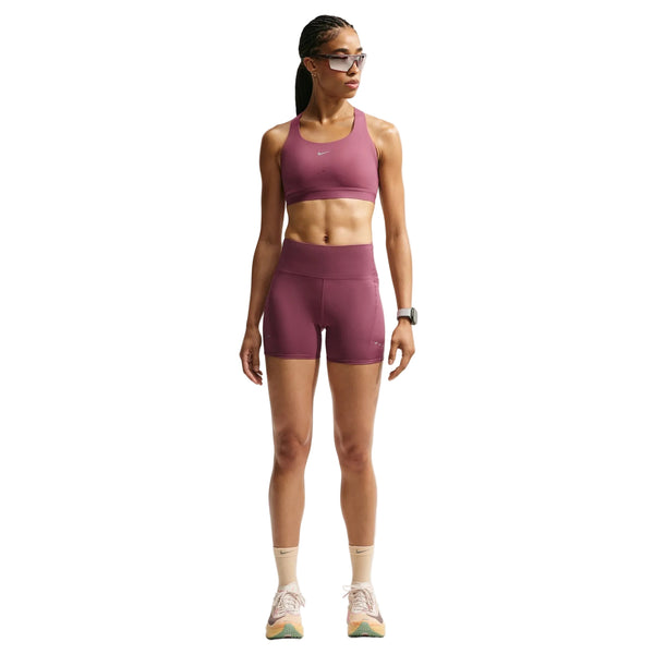 En person står med ansigtet vendt fremad foran en hvid baggrund og har en Nike High-Support Lightly Lined Sports Bra til kvinder i farven Pink Smoke/Reflective Silver på sammen med matchende shorts, hvide sokker og sneakers. Personen har flettet hår, solbriller og et smartwatch på, og armene hænger afslappet ned langs siden.