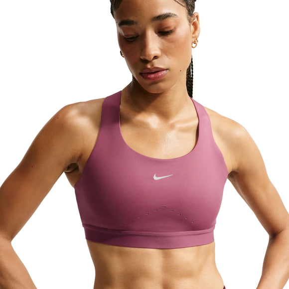 En kvinde har en Nike High-Support Lightly Lined Sports Bra i farven Pink Smoke/Reflective Silver på, med et lille hvidt Nike-logo i midten. BH’en er fremstillet med Dri-FIT-teknologi, der holder dig kølig og tør, og er designet til at give god støtte. Hun kigger let nedad, og hendes hår er sat op i fletninger bagpå.