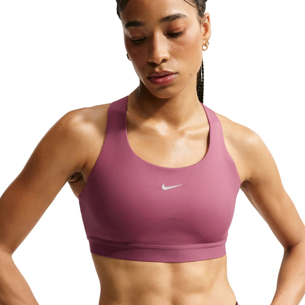 En kvinde har en Nike High-Support Lightly Lined Sports Bra i farven Pink Smoke/Reflective Silver på, med et lille hvidt Nike-logo i midten. BH’en er fremstillet med Dri-FIT-teknologi, der holder dig kølig og tør, og er designet til at give god støtte. Hun kigger let nedad, og hendes hår er sat op i fletninger bagpå.