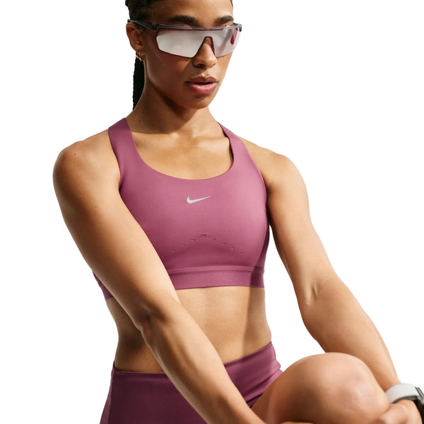 En person, der bærer Nike High-Support Lightly Lined Sports Bra til kvinder i farven Pink Smoke/Reflective Silver, kombineret med matchende leggings med Dri-FIT-teknologi, store solbriller og et smartwatch, holder det ene knæ løftet, som om vedkommende strækker ud eller træner, foran en ensfarvet hvid baggrund.