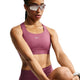 En person, der bærer Nike High-Support Lightly Lined Sports Bra til kvinder i farven Pink Smoke/Reflective Silver, kombineret med matchende leggings med Dri-FIT-teknologi, store solbriller og et smartwatch, holder det ene knæ løftet, som om vedkommende strækker ud eller træner, foran en ensfarvet hvid baggrund.