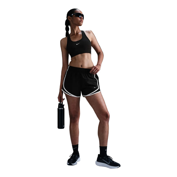 En kvinde med en fletning poserer foran en hvid baggrund iført en sort sports-BH, Nike Tempo Mid-Rise løbeshorts med indvendig underbuks til kvinder i farvekombinationen sort/sort/hvid/reflekterende sølv, sorte sokker og sneakers samt solbriller, og hun holder en vandflaske i hånden.