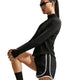 En person har sorte solbriller på, en sort sportstrøje med lange ærmer og Nike Tempo Mid-Rise løbeshorts med indvendig underbuks (Dame, Sort/Sort/Hvid/Reflekterende sølv) og står foran en hvid baggrund, mens vedkommende strækker sig ved at løfte det ene knæ.