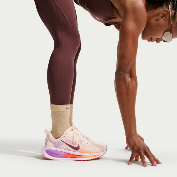 En person i bordeauxfarvet sportstøj og beige sokker bøjer sig forover med en hånd på jorden, iført Nike Vomero 18 Dame i Chalk/Tattoo-Pink Foam-Bright Violet, vist mod en almindelig hvid baggrund.