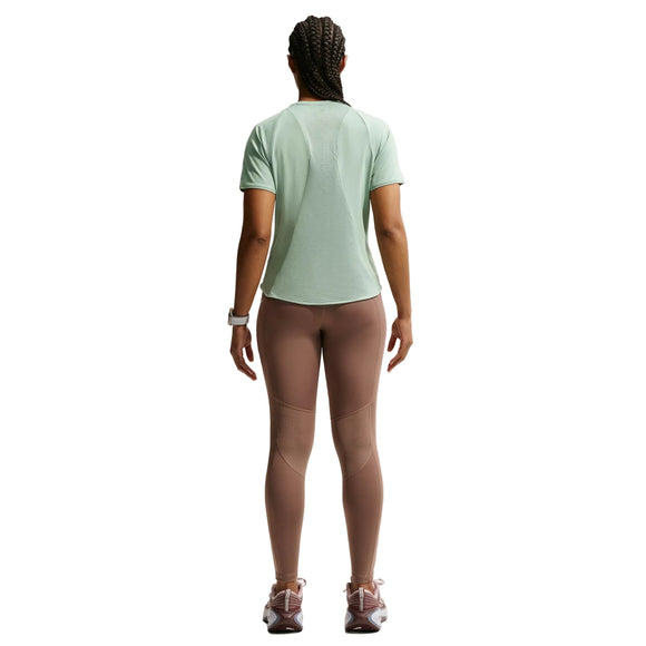 En person med flettet hår kigger væk og er iført en lysegrøn Nike Swift SS Løbetop til kvinder i Steam/Reflective Silver, lilla leggings, pink sneakers og et hvidt armbåndsur på en almindelig hvid baggrund.