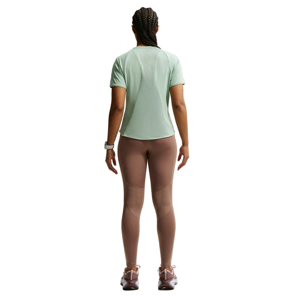 En person med flettet hår kigger væk og er iført en lysegrøn Nike Swift SS Løbetop til kvinder i Steam/Reflective Silver, lilla leggings, pink sneakers og et hvidt armbåndsur på en almindelig hvid baggrund.
