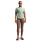 En kvinde står med ansigtet fremad iført en lysegrøn Nike Swift SS Running Top, Dame, Steam/Reflective Silv, taupefarvede leggings, lilla sportssko, solbriller og et ur. Den åndbare løbetop fremhæver komfort og stil mod den hvide baggrund.