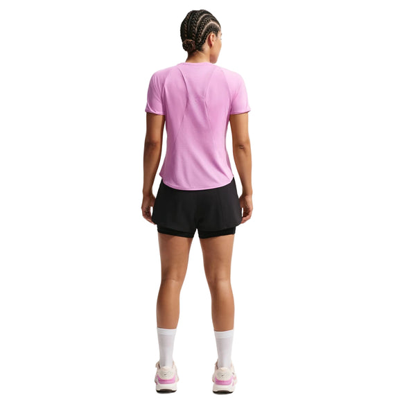 En person med flettet hår står med ryggen til iført en lyslilla Nike Swift SS Løbetop til kvinder (Lt Magenta/Reflective Silver), sorte shorts, hvide sokker og pink-hvide sneakers - og poserer mod en almindelig hvid baggrund.