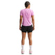 En person med flettet hår står med ryggen til iført en lyslilla Nike Swift SS Løbetop til kvinder (Lt Magenta/Reflective Silver), sorte shorts, hvide sokker og pink-hvide sneakers - og poserer mod en almindelig hvid baggrund.