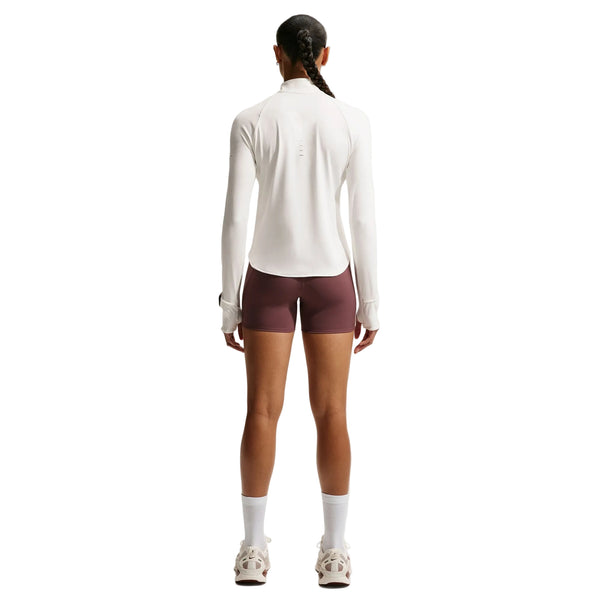 En person står med ryggen til og har en Nike Swift UV Half Zip-løbetrøje til kvinder i farven Sail/Reflective Silver på, kombineret med burgunderfarvede shorts, hvide sokker og hvide sneakers. Håret er sat op i en fletning, og billedet er taget foran en ensfarvet hvid baggrund.