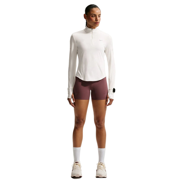 En person står med ansigtet vendt fremad iført en Nike Swift UV Half Zip-løbetrøje til kvinder i farven Sail/Reflective Silver med lange ærmer, lilla sportsshorts, hvide sokker og beige sneakers foran en ensfarvet hvid baggrund. Dette sæt kombinerer komfortabel stil med optimal UV-beskyttelse til aktive dage.