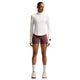 En person står med ansigtet vendt fremad iført en Nike Swift UV Half Zip-løbetrøje til kvinder i farven Sail/Reflective Silver med lange ærmer, lilla sportsshorts, hvide sokker og beige sneakers foran en ensfarvet hvid baggrund. Dette sæt kombinerer komfortabel stil med optimal UV-beskyttelse til aktive dage.